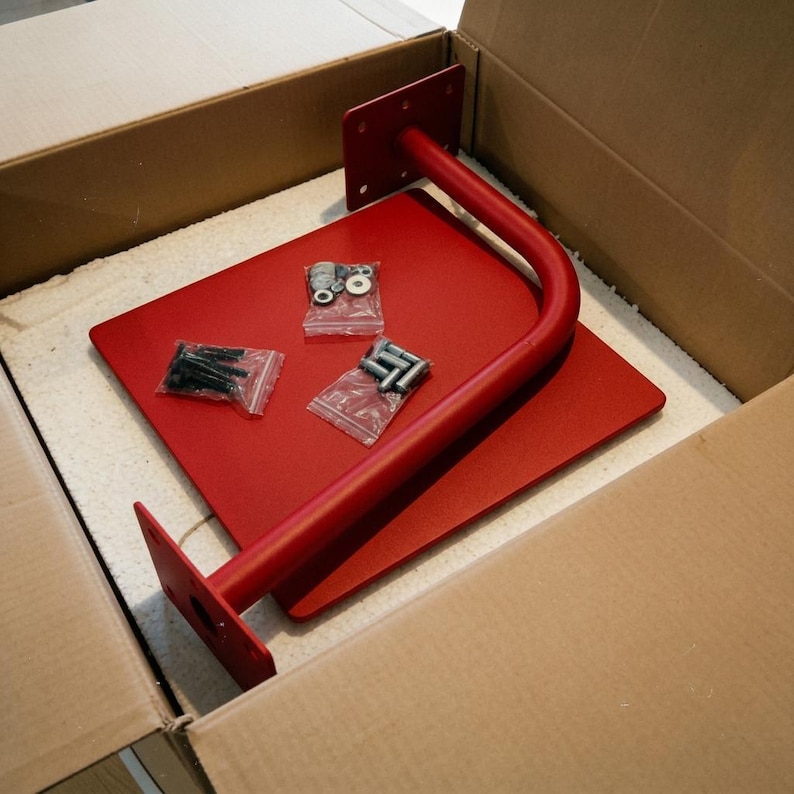 May include: A red metal wall-mounted bracket featuring a flat square base plate and a curved support arm with mounting flanges. Three small clear plastic bags containing silver-colored metal screws, bolts, and washers are placed on the flat surface.