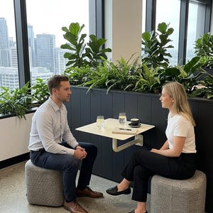 May include: Two individuals sit on gray fabric ottomans facing each other at a small, square, off-white wall-mounted table. On the table are two glasses of water, a black coffee cup on a saucer, and a notebook with a black pen resting on top.