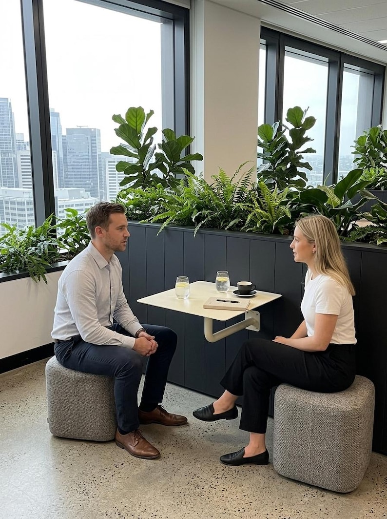 May include: Two individuals sit on gray fabric ottomans facing each other at a small, square, off-white wall-mounted table. On the table are two glasses of water, a black coffee cup on a saucer, and a notebook with a black pen resting on top.