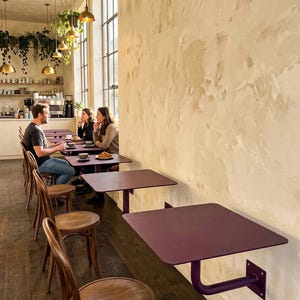 May include: A series of wall-mounted, square, dark purple metal tables with rounded corners, each supported by a single matching purple L-shaped bracket. The tables are arranged in a row along a textured, light-colored wall in a cafe setting.