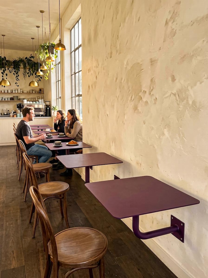 May include: A series of wall-mounted, square, dark purple metal tables with rounded corners, each supported by a single matching purple L-shaped bracket. The tables are arranged in a row along a textured, light-colored wall in a cafe setting.