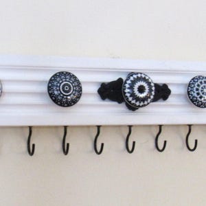 May include: White wooden wall mounted key rack with nine black metal hooks. The rack is decorated with four black and white ceramic knobs with intricate patterns.