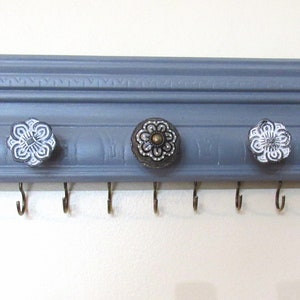 May include: Gray wooden key holder with eight hooks and five decorative knobs. The knobs are white with black floral designs and have a gold center.