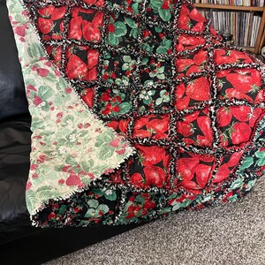 May include: A handmade rag quilt featuring a patchwork design of red strawberries and green leaves on a black background. The quilt has a folded-over corner revealing a light green backing fabric with a smaller strawberry and vine print pattern.