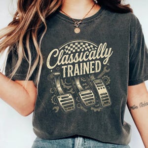 Może przedstawiać: Ciemnoszara koszulka z napisem "Classically Trained" nad grafiką trzech pedałów samochodowych, kół zębatych i kluczy. Projekt zawiera wzór w szachownicę. Koszulka jest w stylu vintage.