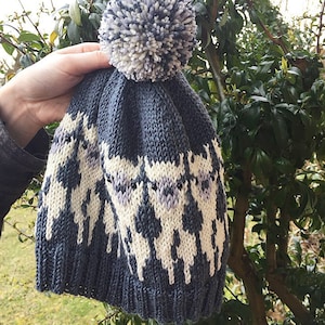 Könnte beinhalten: Eine graue Strickmütze mit einem weißen und lila Alpaka-Muster und einem weißen und grauen Bommel oben.