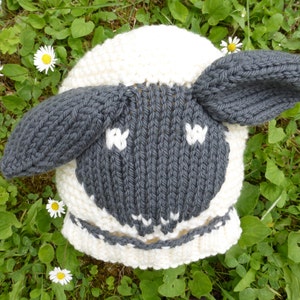May include: A white and gray knitted hat shaped like a sheep with black eyes and a white nose.