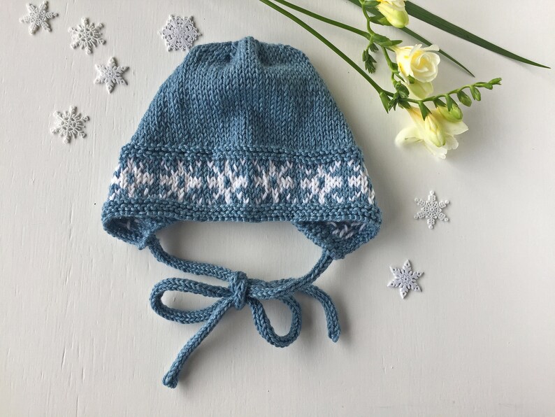 May include: A blue knitted baby hat with a white snowflake pattern around the brim. The hat has a tie that can be tied under the chin.
