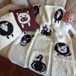 May include: Cream-colored knitted baby blanket and pillows with farm animal designs. The blanket features a grid pattern with black and white sheep, pig, and duck motifs. Two pillows with sheep and pig designs are also visible.