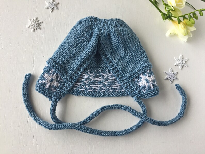 May include: A blue knitted baby hat with white snowflake designs and matching ties. The hat is folded down and the ties are tied together.