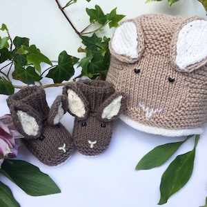 May include: A set of knitted baby booties and a hat, both featuring bunny ears and faces. The booties are brown with white ears and the hat is beige with white ears.
