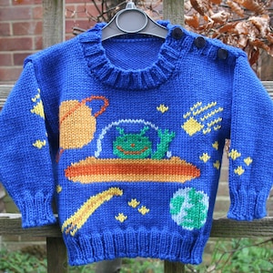 Peut inclure: Un pull en tricot bleu sur le thème de l'espace. Le pull présente un extraterrestre vert dans une soucoupe volante, une planète, une comète et des étoiles.