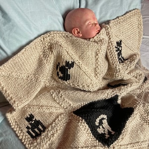 Puede incluir: Una manta de bebé de punto beige con diseños de animales negros. La manta está doblada y un bebé está durmiendo encima.