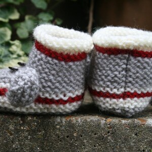 May include: A pair of gray knitted baby booties with white and red stripes. The booties have a textured knit pattern and are made with soft yarn.