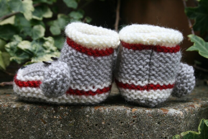 May include: A pair of gray knitted baby booties with white and red stripes. The booties have a textured knit pattern and are made with soft yarn.