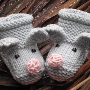 May include: Two gray knitted pig slippers with pink snouts and black eyes. The slippers are sitting on a bed of brown dried leaves.