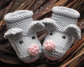 May include: Two gray knitted pig slippers with pink snouts and black eyes. The slippers are sitting on a bed of brown dried leaves.