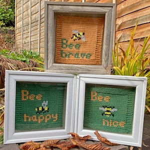 May include: Three framed knitted squares with bee and text designs. The top square is orange with a bee and the text "Bee Brave". The middle square is green with a bee and the text "Bee Happy". The bottom square is green with a bee and the text "Bee Nice".