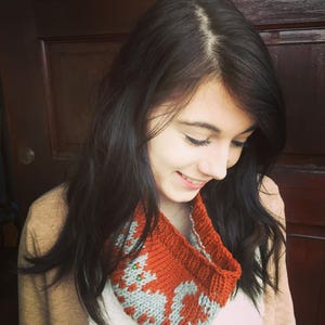 May include: A knitted cowl scarf in shades of rust orange and gray. The scarf features a geometric pattern with a repeating design. The cowl is worn around the neck and is made of a soft, warm material, perfect for cold weather.