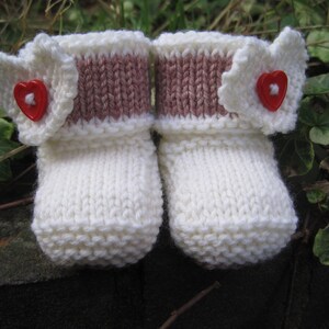 May include: A pair of white knitted baby booties with pink and white trim and red heart buttons.