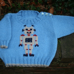 May include: A light blue knitted sweater with a robot design in the center. The robot is white with red, blue, and black details. The sweater has a ribbed collar and cuffs.