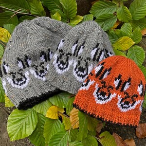 Puede incluir: Tres gorros de punto con un diseño gráfico en blanco y negro. Un gorro es gris, otro es blanco y otro es naranja.