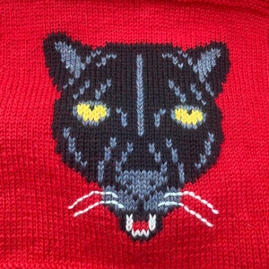 Può includere: Un viso di pantera nera lavorato a maglia su un maglione rosso. La pantera ha occhi gialli e baffi bianchi.