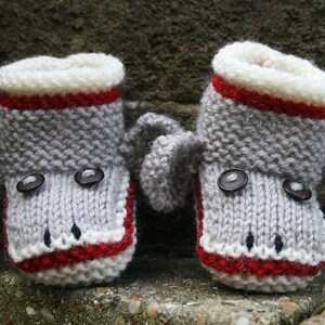 May include: A pair of gray knitted baby booties with red and white trim. The booties have button eyes and a gray knitted elephant ear on each side.