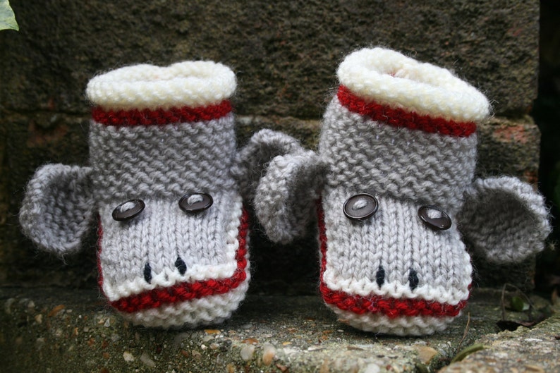 May include: A pair of gray knitted baby booties with red and white trim. The booties have button eyes and a gray knitted elephant ear on each side.