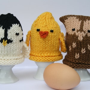 May include: Three knitted egg cosies, one penguin, one chick, and one owl, each sitting on a white egg cup.