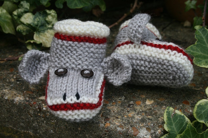 May include: A pair of gray knitted baby booties with red and white stripes, featuring a monkey face with black button eyes.