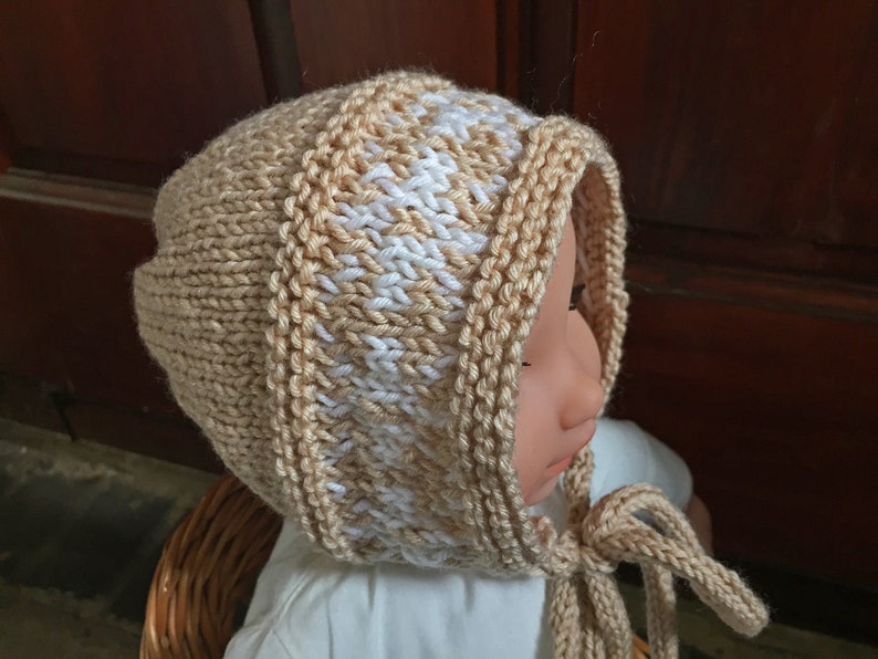 May include: A knitted baby bonnet with a beige and white striped pattern. The bonnet has a tie that can be tied under the chin.
