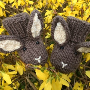 May include: Two knitted brown bunny rabbit baby booties with white ears. The booties are sitting on a bed of yellow flowers.