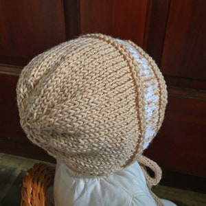 May include: A knitted baby bonnet in beige with a white trim. The bonnet has a tie at the back.