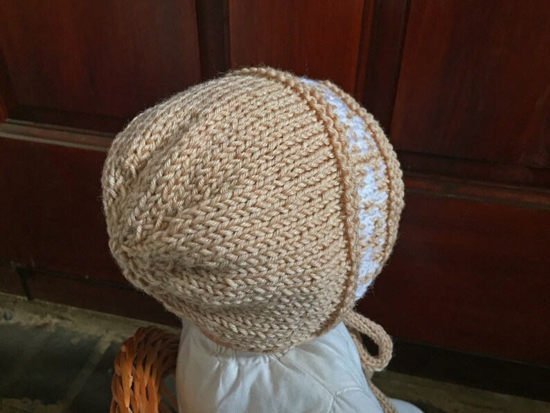 May include: A knitted baby bonnet in beige with a white trim. The bonnet has a tie at the back.
