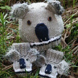 Könnte beinhalten: Eine graue gestrickte Koala-Mütze und passende Fäustlinge. Die Mütze hat einen weißen Rand und schwarze Augen und Nase. Die Fäustlinge haben einen weißen Rand und schwarze Augen und Nasen.