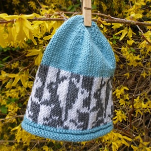 May include: A blue and gray knitted hat with a geometric pattern. The hat is hanging from a wooden clothespin against a background of yellow flowers.