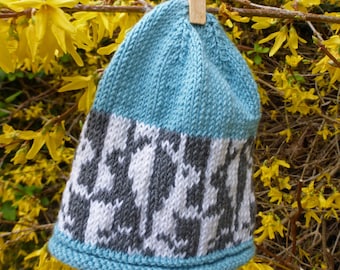Könnte beinhalten: Eine blaue und graue Strickmütze mit geometrischem Muster. Die Mütze hängt an einer Holzklammer vor einem Hintergrund aus gelben Blumen.
