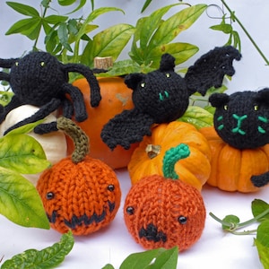 May include: Four knitted Halloween decorations: a black spider, a black bat, a black cat, and an orange pumpkin. The decorations are sitting on top of orange pumpkins.