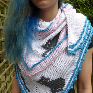 Peut inclure: Un châle tricoté blanc, rose et bleu avec un motif géométrique. Le châle a une forme triangulaire avec une bordure bleue festonnée. Le motif comprend un motif géométrique gris. Le châle est porté autour du cou.