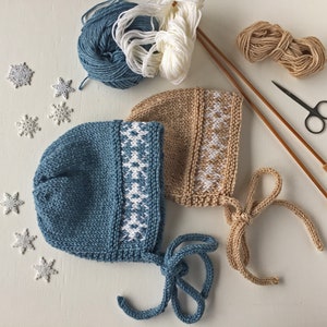May include: Two knitted baby hats with white snowflake patterns. One hat is blue and the other is beige. The hats have ties that can be tied under the chin.