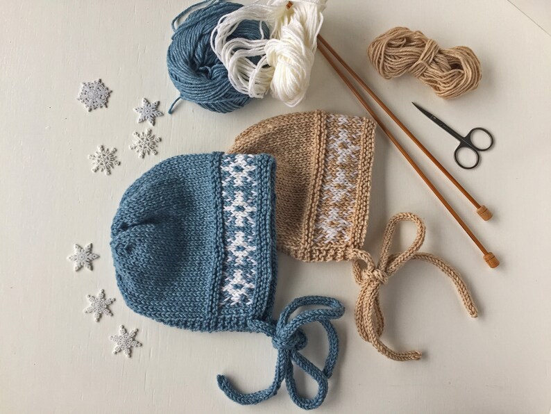 May include: Two knitted baby hats with white snowflake patterns. One hat is blue and the other is beige. The hats have ties that can be tied under the chin.