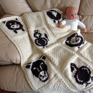May include: A white knitted baby blanket with brown animal motifs. The blanket features a sheep, a cat, a dog, and a bird. The blanket is on a couch.