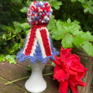 May include: A hand-knitted hat with a red, white, and blue color scheme, featuring a pom-pom on top. The hat has a scalloped edge and is displayed on a white ceramic stand. A vibrant red rose is in the foreground.
