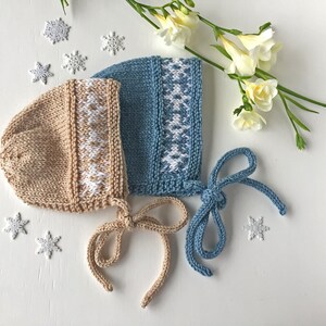 May include: Two knitted baby bonnets with white snowflake patterns. One bonnet is beige and the other is blue.