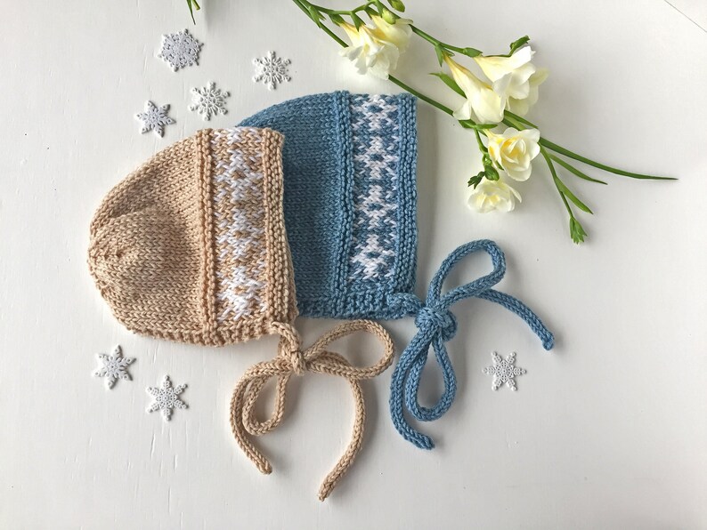 May include: Two knitted baby bonnets with white snowflake patterns. One bonnet is beige and the other is blue.