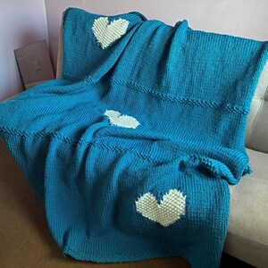 May include: A teal knitted blanket with three white heart appliqués. The blanket is draped over a sofa, showcasing its textured knit and the contrasting white hearts. The blanket's design is simple and cozy, perfect for home decor.
