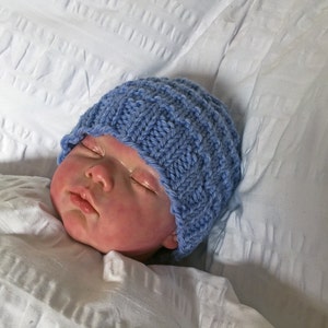 May include: A newborn baby wearing a blue knitted hat is sleeping on a white sheet.