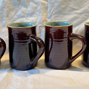 May include: A set of four cylindrical ceramic mugs featuring a glossy dark brown exterior with horizontal ridged textures and a contrasting light sage green interior glaze. Each mug includes a sturdy, curved handle attached to the side.