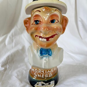 May include: A vintage ceramic bottle shaped like a smiling cartoon man wearing a hat and bow tie. The bottle is tan, with blue eyes and a red mouth. The text "Mortimer Snerd" is printed on the base.